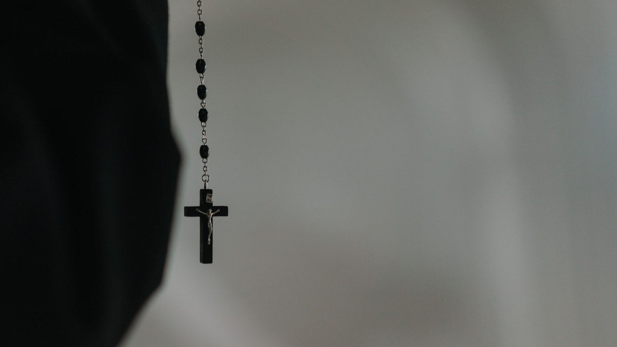 Chapelet a grains noirs monte sur une fine chaine metallique sombre, crucifix noir mat, suspendu devant un fond gris