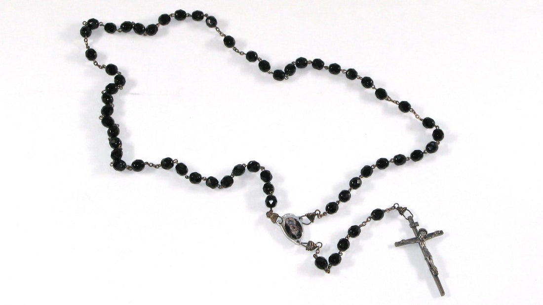 Chapelet ancien en grains de verre noir monte sur chaine, avec sa croix, sa medaille de jonction ovale et sa boucle de cinq dizaines, photographie a plat sur fond clair.