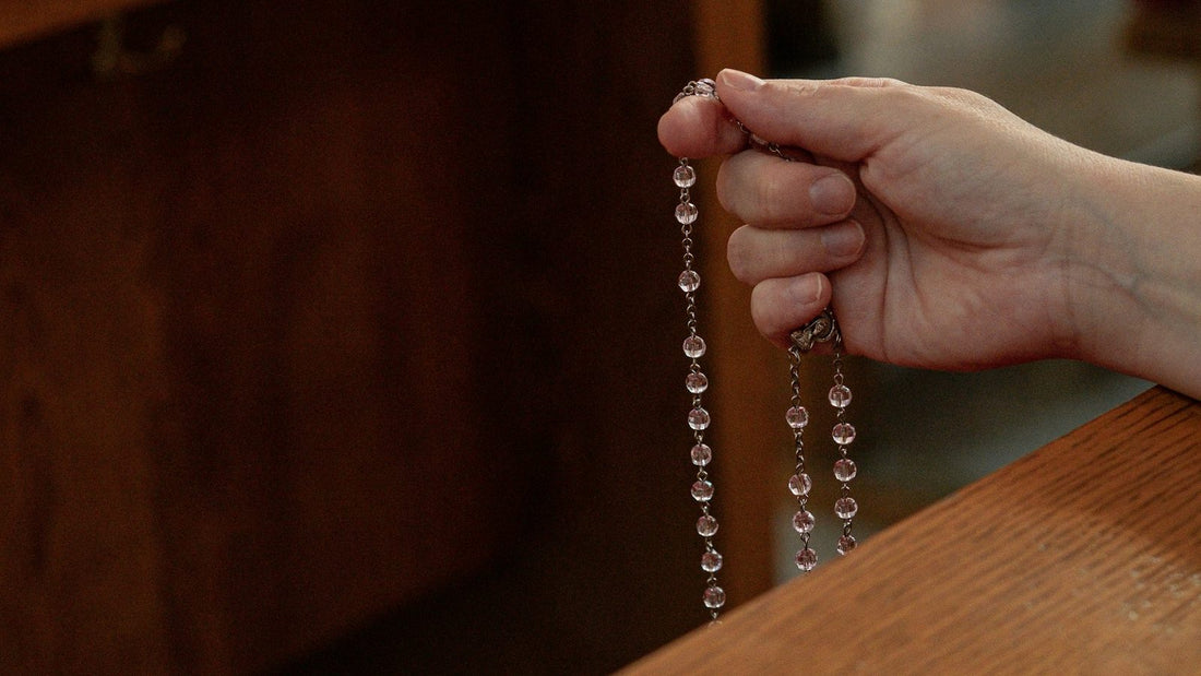 Une main tient un chapelet de grains de cristal au bord d'un banc d'eglise, le reste du brin pendant librement.