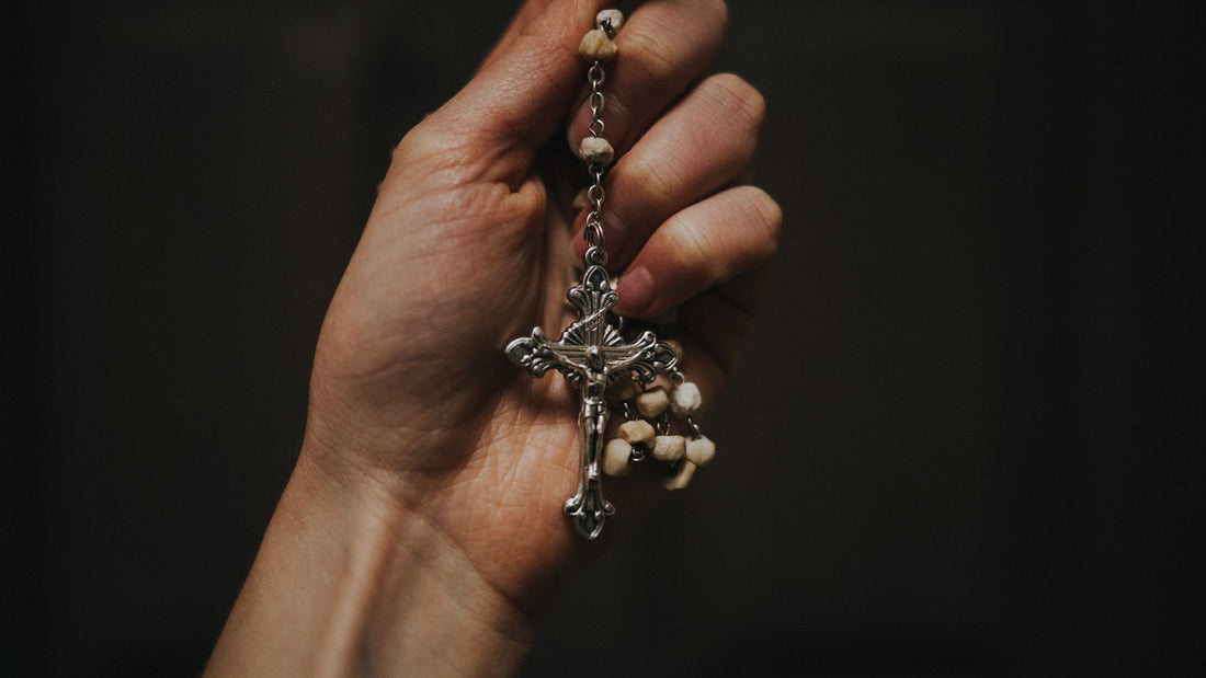Une main tient un dizainier de poignet par son crucifix, les grains clairs rassemblés dans la paume, sur fond sombre