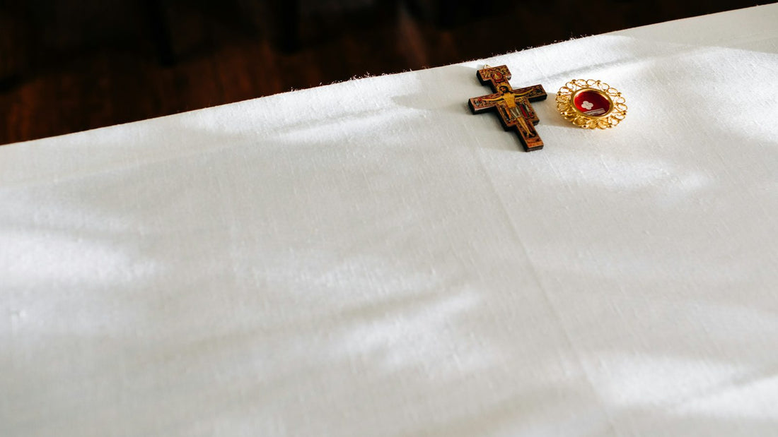Croix de bois peinte et petit reliquaire dore poses sur la nappe d'autel en lin blanc, sous une lumiere rasante.