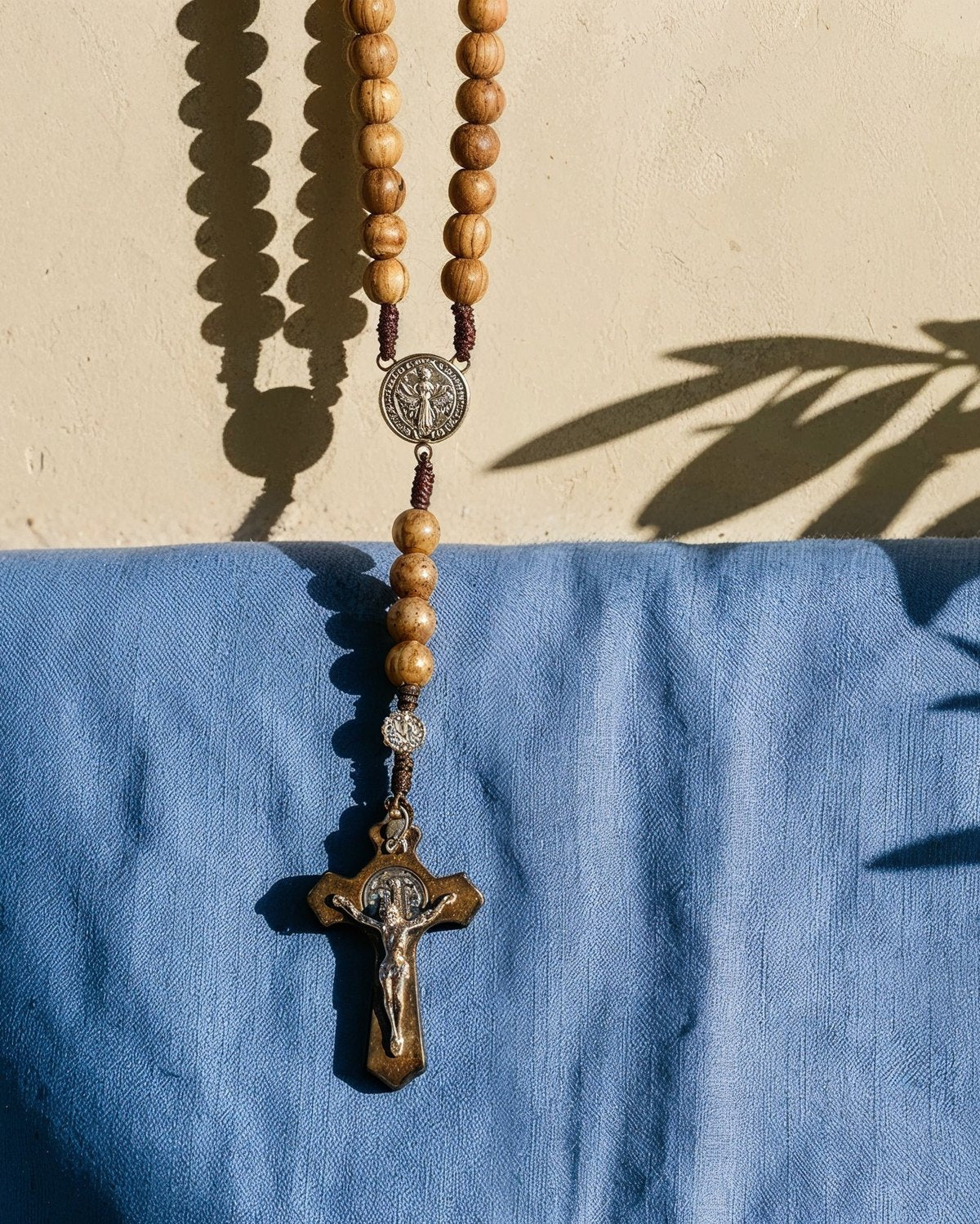 Un chapelet en bois de saint Benoît suspendu au-dessus d'un lin bleu, l'ombre d'un olivier sur le mur.